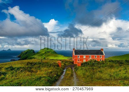 A Red House In The Ireland Countryside. The Cloudy Sky Contrasting With The Green Of The Huge Fields
