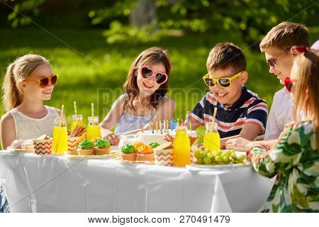 holidays, childhood and celebration concept - happy kids with candles on birthday cake sitting at table at summer garden party