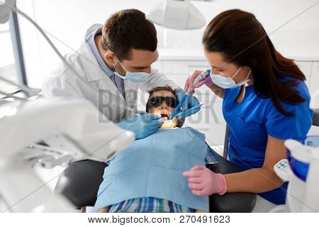 medicine, dentistry and healthcare concept - dentist and assistant with dental drill and saliva ejector treating kid patient teeth at dental clinic