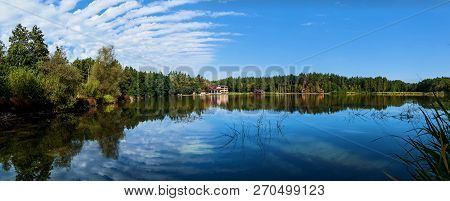 Wooden House Near The Lake With Clear Water. The Pond Reflects The Dense Green Forest And Sky With W