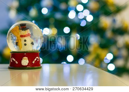 Snowman Snow Globe In From Of Defocused Christmas Tree With Gold Decorations And White Lights.