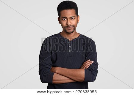 Photo Of Doubtful Serious Dark Skinned Man Keeps Hands Crossed, Hears Some Information, Dressed In C