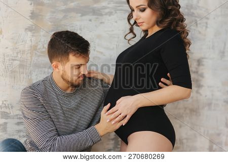 Happy Man Put His Handr On The Belly Of His Pregnant Wife. Young Family In Studio. Couple Wait For A