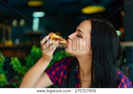 Beautiful Brunette Girl In T-shirt Eating Pizza At Restaurant. A Pretty Girl Feels Happy And Enjoys 