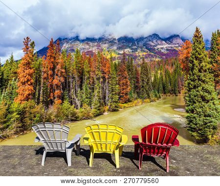 Scenic autumn in the Rockies of Canada. Three deck chairs stand over the forest and river. Thunderclouds swirl in the sky. Concept of ecological, active and photo-tourism