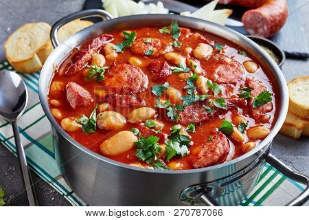 Hearty Beans Stew With Sausages, Herbs And Spices In Tomato Sauce In A Metal Casserole On A Concrete