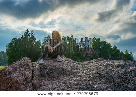 Pretty Young Lady With Red Hair Sitting On Stone Near Bay Shore In Summer Evening. Tourist On The Be