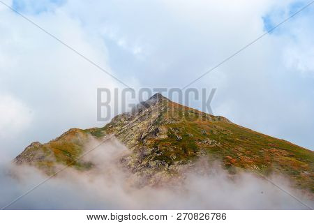 Cone-shaped Rocky Mountain Peak With Multi-colored Vegetation Visible In The Lumen Of The Clouds