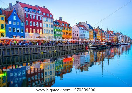 People Walking And Sitting In Restaurants At Illuminated Nyhavn Embankment By Canal With Moored Boat