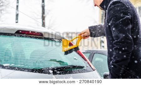 Young Man In Black Coat Cleans His Car With Yellow Brush During Snowfall. Winter Inclement Weather.