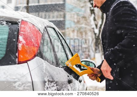 Young Man In Black Coat Cleans His Car With Yellow Brush During Snowfall. Winter Inclement Weather.