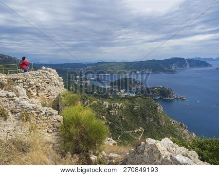 Older Woman In Red Jacket Looking At View Of The Paleokastritsa Bay And Monastery From The View Poin