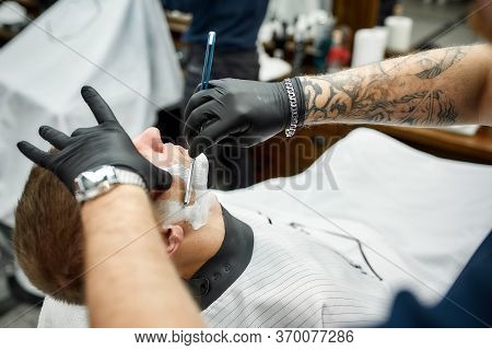 Traditional Shave. Cropped Photo Of Barber In Black Gloves With Tattooed Arm Shaving Client With Dan