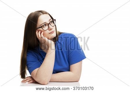 A Pensive Girl In Glasses And A Blue T-shirt Sits Propping Her Head On Her Hand. Isolated On A White