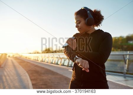 Choosing Music Playlist For Run. Sporty African Woman Wearing Headphones, Using Smatphone, Standing 