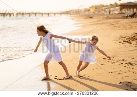 Girl Children In White Dresses Playing On The Beach. Older Sister Pulls The Younger Into The Water. 