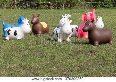 A Herd Of Inflatable Hopper Toys In The Field. Green With Yellow Flowers Pasture, Trees In The Backg