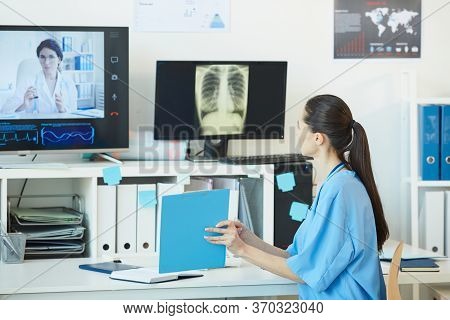 Back View At Young Female Medic Talking To Doctor By Video Call And Showing Documents While Sitting 