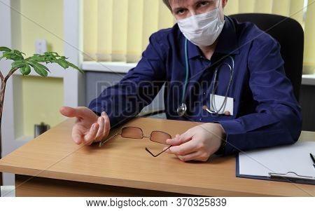 A Doctor In A Medical Mask On His Face Explains Something To The Patient. He Sits At The Table And G
