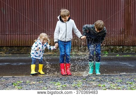 Three Children, Toddler Girl And Two Kids Boys Wearing Red, Yellow And Green Rain Boots And Walking 