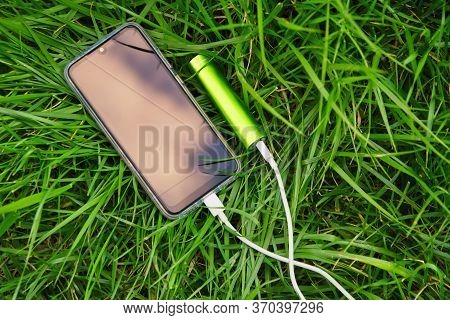Smartphone And External Power Bank Lie On Green Grass In The Park While Charging