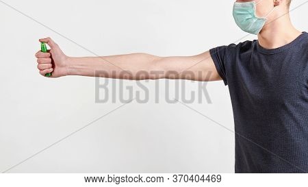 Man In Dark Shirt And Protective Medical Mask Is Spraying Hand Sanitizer. Light Background, Closeup.