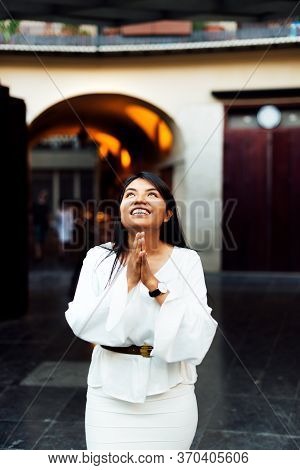 Latin Brunette Woman Dressed In White Smiling Looking Up At The Sky With Her Hands Together In A Pra