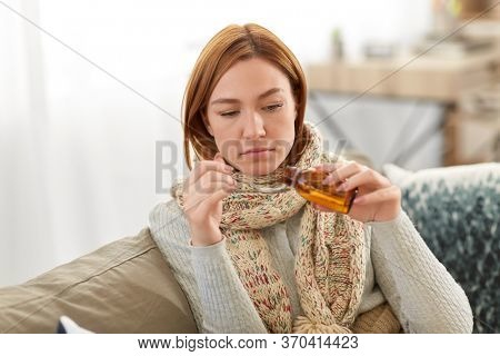 people, health and cold concept - sad sick woman in blanket and scarf pouring cough syrup or antipyretic medicine at home