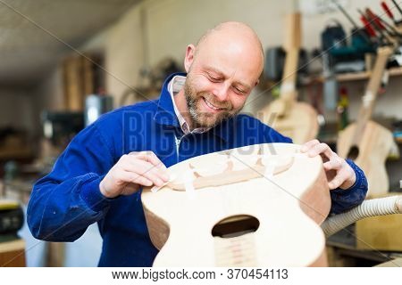Portrait Of Positive Smiling Guitar Maker Holding Unfinished Guitar At Workshop