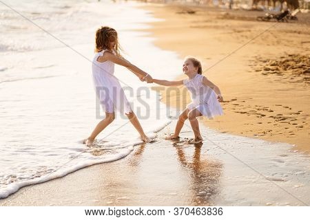 Girl Children In White Dresses Playing On The Beach. Older Sister Pulls The Younger Into The Water. 