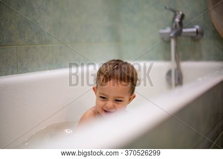 Cute Caucasian Baby Take A Bath, Playi In Bath And Smiles Looking At Water Splashes, In The Backgrou