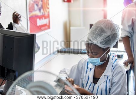 African woman working at a butchery shop,  taking cash at the point of sale, using a mask for protection and a hairnet 