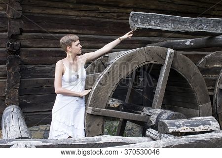 A Short-haired Girl In A White Linen Gown, Reaches Out To A Wooden Frame With A Wheel, An Old Water 