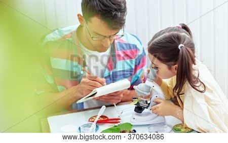 Child Looks Through Microscope While Doing Science Experiments In The Laboratory With Her Father. Da