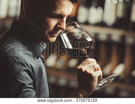 Sommelier Smelling Flavor Of Red Wine In Bokal On Background Of Shelves With Bottles In Cellar. Male