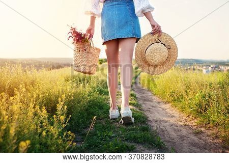 Young Woman With Straw Hat And Bag Of Wildflowers Walks Along Path Into Distance At Sunset