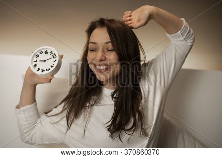 Young Attractive Female With Dark Hair In Bed Wakes Up From An Alarm Clock, Stretches And Broadly Sm