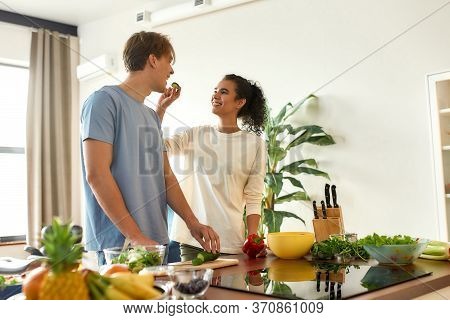 Young Man Cutting Vegetables While Woman Feeding Him A Slice Of Cucumber. Vegetarians Preparing Heal