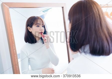 Asian Woman Choosing Make Up With Cosmatic Applying Lipstick And Smiling Happy In Front Mirror At Ho