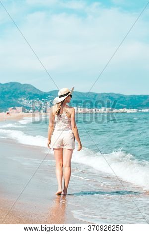 Back View Of A Woman Walking Towards The Sea. Woman Alone Exploring Mediterranean Sea Beach During S