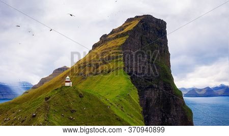 Small White Kallur Lighthouse Located Near Huge Cliffs On Island Of Kalsoy With Seagulls Flying. Kal