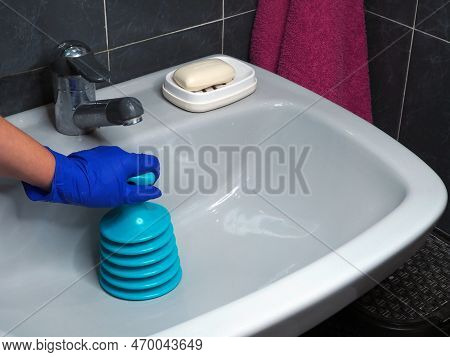 White Ceramic Sink And Plunger In Hand Clears Blockage. Plumbing Tool For Mechanical Cleaning Of Blo