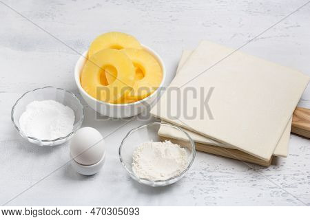 Baking Ingredients: Puff Pastry, Pineapple Rings, Flour, Egg And Powdered Sugar On A Light Gray Back