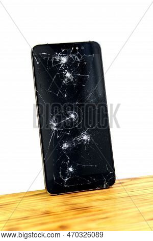 A Broken Cell Phone Stands On A Wooden Table Tilted Against A White Isolated Wall.