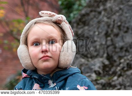 Autumn Portrait Of A Girl 7-8 Years Old, With Wide Open Surprised Eyes, In Warm Fluffy Headphones