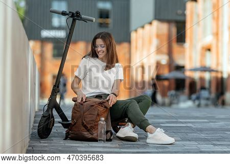 Young Female Sitting On Urban Stairs And Browsing Smart Watch On Electric Scooter