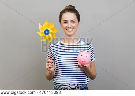 Portrait Of Woman Wearing Striped T-shirt Holding Origami Hand Mill On Stick And Piggy Bank, Looking