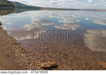 Lake Surrounded By A Rocky Shore, Mountains With Forest On Background.reflection Of White Clouds In 