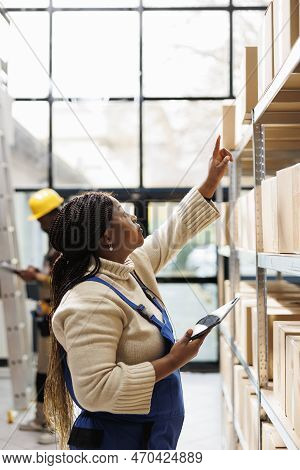Warehouse Worker Reaching For Parcel At Cardboard Boxes Shelf And Doing Goods Inventory With Tablet 