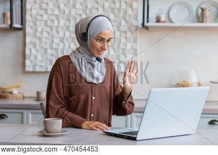 Woman In Hijab Working Remotely From Home Sitting In Kitchen Using Laptop And Headset For Video Call